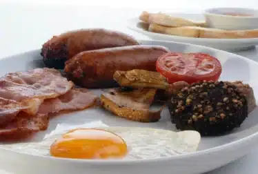 A breakfast plate with fried egg, bacon, sausages, grilled tomato, mushrooms, black pudding, and toast with a small bowl of jam in the background. shoreline
