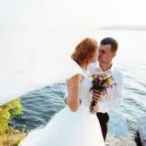A bride and groom stand together by a lake, with the bride holding a bouquet and her veil blowing in the wind.