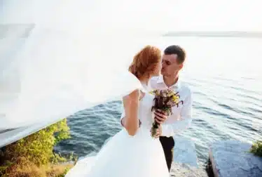 A bride and groom stand together by a lake, with the bride holding a bouquet and her veil blowing in the wind.
