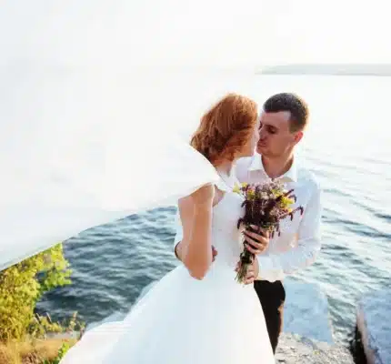 A bride and groom stand together by a lake, with the bride holding a bouquet and her veil blowing in the wind.