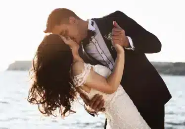 A couple in formal wedding attire shares a kiss while dancing on a beach with the ocean and bright sunlight in the background.