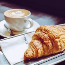 A cup of coffee with latte art sits on a saucer next to a croissant on a metal tray with a Fonté brandednnapkin and knife.