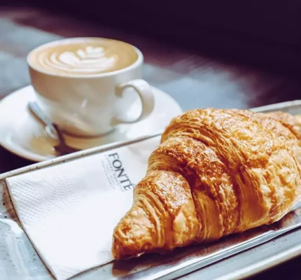 A cup of coffee with latte art sits on a saucer next to a croissant on a metal tray with a Fonté brandednnapkin and knife.