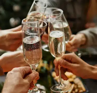 A group of people clink champagne glasses in a toast, with food and a decorated tree visible in the background.
