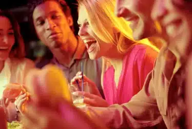 A group of people sit closely together at a table, smiling and laughing; one woman holds a drink with a straw.