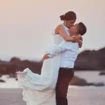 A man in a white shirt lifts a woman in a white dress on a beach at sunset, with rocks and waves in the background.