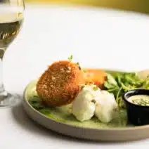 A plate of breaded fried cakes, poached eggs, green salad, a small cup of sauce, and a glass of white wine on a white tablecloth.