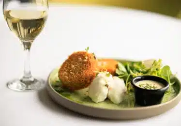 A plate of breaded fried cakes, poached eggs, green salad, a small cup of sauce, and a glass of white wine on a white tablecloth.