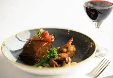 A plate with braised meat, vegetables, and sauce is next to a glass of red wine on a white tablecloth.