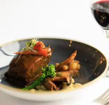 A plate with braised meat, vegetables, and sauce is next to a glass of red wine on a white tablecloth.