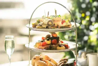 A three-tiered stand with assorted finger sandwiches, pastries, fruits, and scones, accompanied by two glasses of sparkling drink on a table. Christmas tree in background