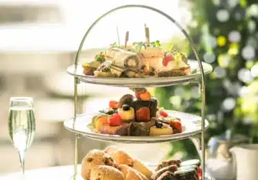 A three-tiered stand with assorted finger sandwiches, pastries, fruits, and scones, accompanied by two glasses of sparkling drink on a table. Christmas tree in background