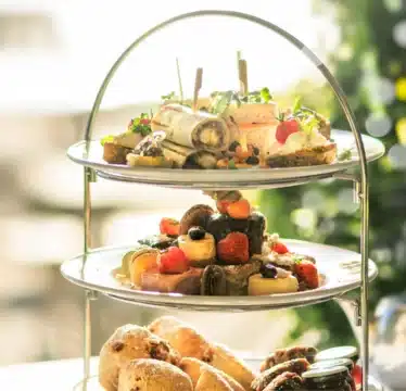 A three-tiered stand with assorted finger sandwiches, pastries, fruits, and scones, accompanied by two glasses of sparkling drink on a table. Christmas tree in background