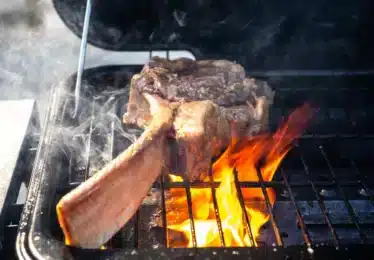 A tomahawk steak is being grilled over open flames on a barbecue, with other pieces of meat cooking on an upper rack.