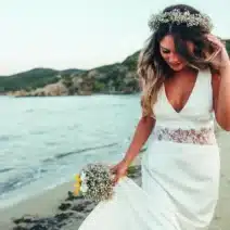 A woman in a white wedding dress and flower crown walks along a sandy beach, holding a bouquet and smiling with the sea and hills in the background.