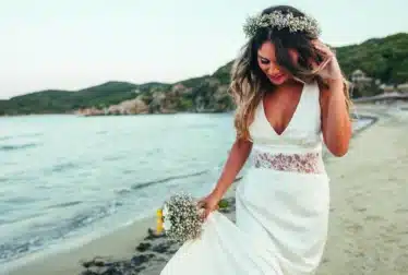 A woman in a white wedding dress and flower crown walks along a sandy beach, holding a bouquet and smiling with the sea and hills in the background.