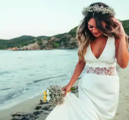 A woman in a white wedding dress and flower crown walks along a sandy beach, holding a bouquet and smiling with the sea and hills in the background.