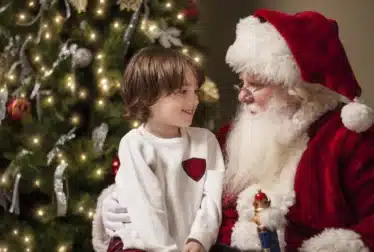 A young boy sits on Santa Claus’s lap, smiling, in front of a decorated Christmas tree with lights and ornaments.