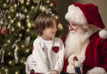 A young boy sits on Santa Claus’s lap, smiling, in front of a decorated Christmas tree with lights and ornaments.