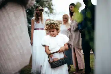A young flower girl in a white dress and floral crown holds a small basket, while several adults and another girl stand in the background outdoors.
