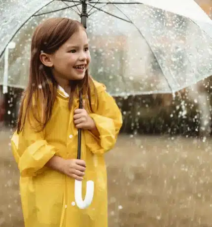 A young girl in a yellow raincoat stands under a clear umbrella while it rains, smiling and looking to the side in an outdoor setting.