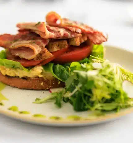 Brown bread with scrambled eggs, tomatoes, bacon and a side salad on a plate on a white table cloth