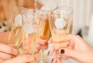 Five hands holding champagne flutes are toasting together, with each glass partially filled with sparkling wine. The background is out of focus.
