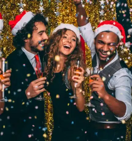 Five people wearing Santa hats celebrate together, holding drinks and smiling, with gold tinsel background and confetti falling.