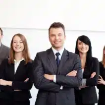 Five professionally dressed people stand in a row, all smiling, with the person in the center slightly forward and arms crossed.