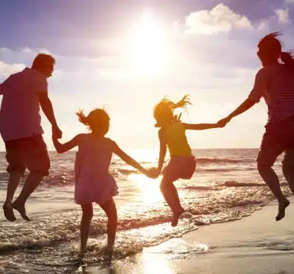 Four people, two adults and two children, are holding hands and jumping on a beach shoreline at sunset with the ocean in the background.