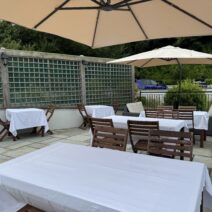 Outdoor patio dining area with wooden tables and chairs covered in white tablecloths, shaded by large cream umbrellas, and surrounded by greenery and a wooden trellis.