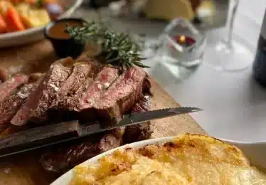 Sliced medium-rare steak on a wooden board with a knife, accompanied by a dish of gratin potatoes and a bottle of wine on a set dining table.