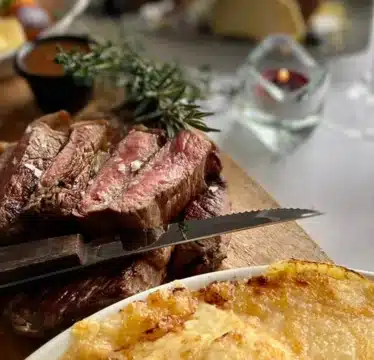 Sliced medium-rare steak on a wooden board with a knife, accompanied by a dish of gratin potatoes and a bottle of wine on a set dining table.