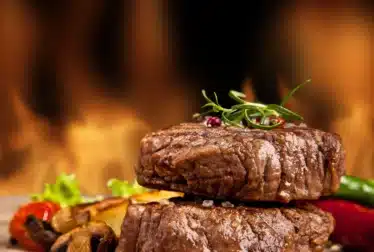 Two grilled steaks stacked on a wooden board, garnished with herbs and peppercorns, with mushrooms, vegetables, and a flame background.