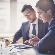 Two men in business suits sit at a desk, discussing documents and using a digital tablet in a bright office setting.