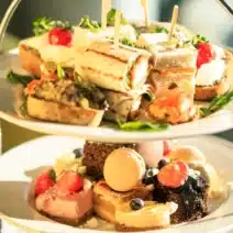 Two-tiered tray with assorted finger sandwiches, pastries, macarons, and fruit, set on a table with a glass of sparkling beverage in the background.