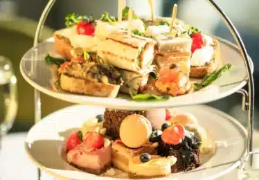 Two-tiered tray with assorted finger sandwiches, pastries, macarons, and fruit, set on a table with a glass of sparkling beverage in the background.