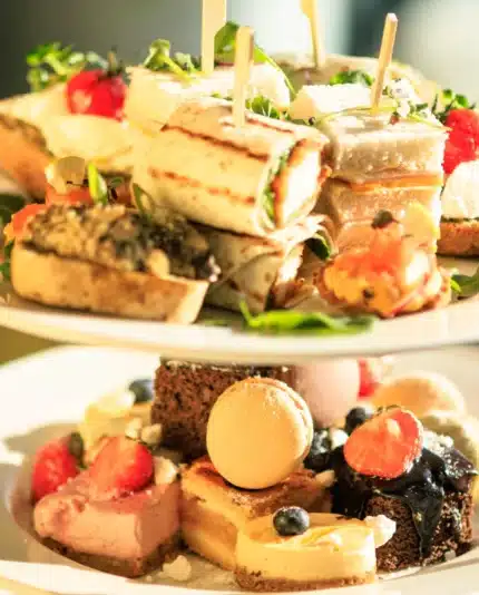 Two-tiered tray with assorted finger sandwiches, pastries, macarons, and fruit, set on a table with a glass of sparkling beverage in the background.