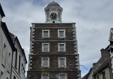 The historic Clock Gate Tower in Youghal, County Cork, standing tall above the narrow street with surrounding colorful and traditional buildings.