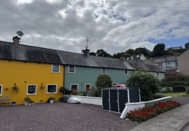 Row of charming colourful cottages in Youghal, County Cork, with bright yellow and green facades, potted plants, and a well-kept garden under a cloudy sky.
