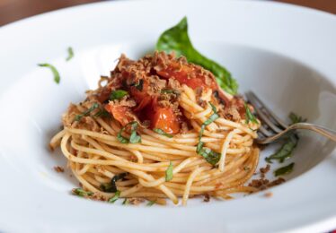 Close-up of freshly prepared spaghetti with tomato sauce, basil, and crispy topping, served in a white bowl with a fork.