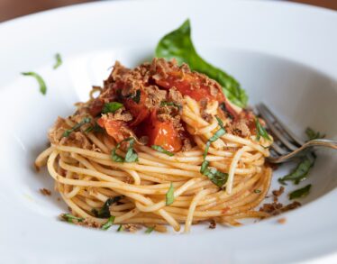 Close-up of freshly prepared spaghetti with tomato sauce, basil, and crispy topping, served in a white bowl with a fork.