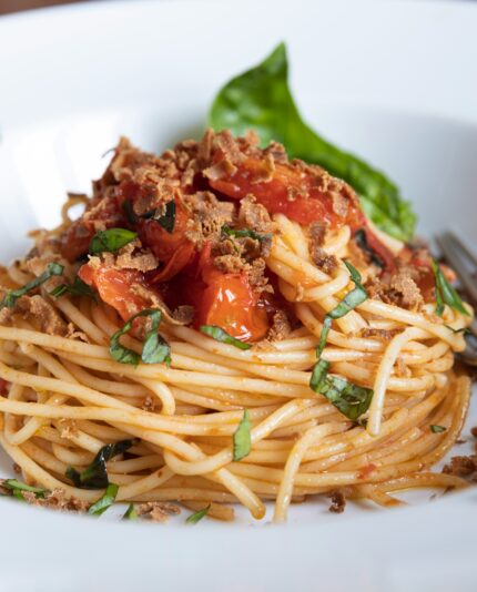 Close-up of freshly prepared spaghetti with tomato sauce, basil, and crispy topping, served in a white bowl with a fork.